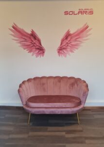 Zdjęcie nadruku ściennego różowych anielskich skrzydeł przed którymi stoi elegancka, różowa sofa.